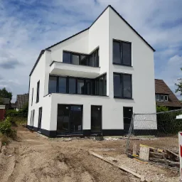 Nowoczesny, dwupiętrowy dom z białą elewacją i czarnymi ramami okiennymi, częściowo wykończony, z widocznym terenem budowy przed budynkiem.