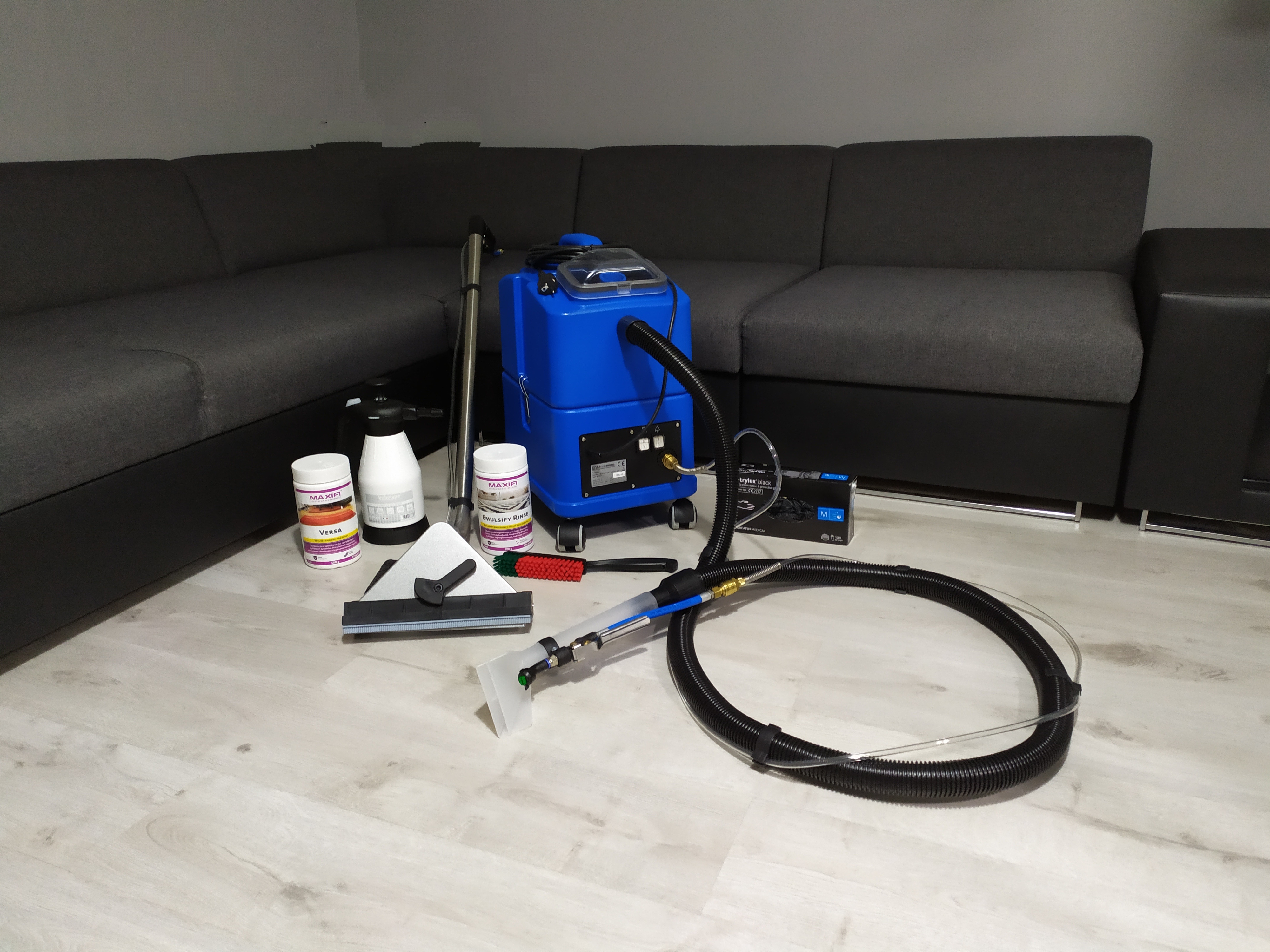 Profesjonalny sprzęt do czyszczenia tapicerki, w tym niebieski ekstraktor, różne końcówki, szczotka i środki czyszczące Maxifi, rozłożony na jasnej podłodze na tle szarej sofy modułowej.