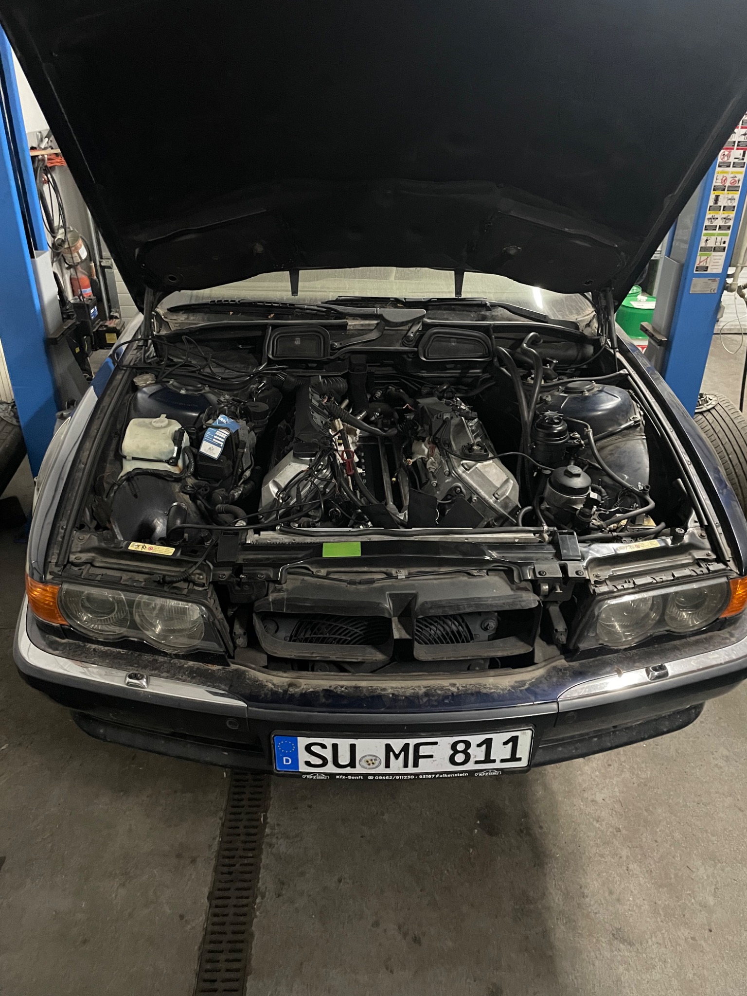 Niebieski samochód z otwartą maską w warsztacie, widoczny silnik w trakcie naprawy, podnośnik warsztatowy w tle.