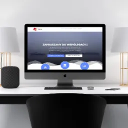 Ekran komputera iMac prezentujący stronę internetową StarTrade z hasłem 'Zapraszamy do współpracy', na białym biurku z lampami i głośnikiem, widok zza czarnego krzesła.