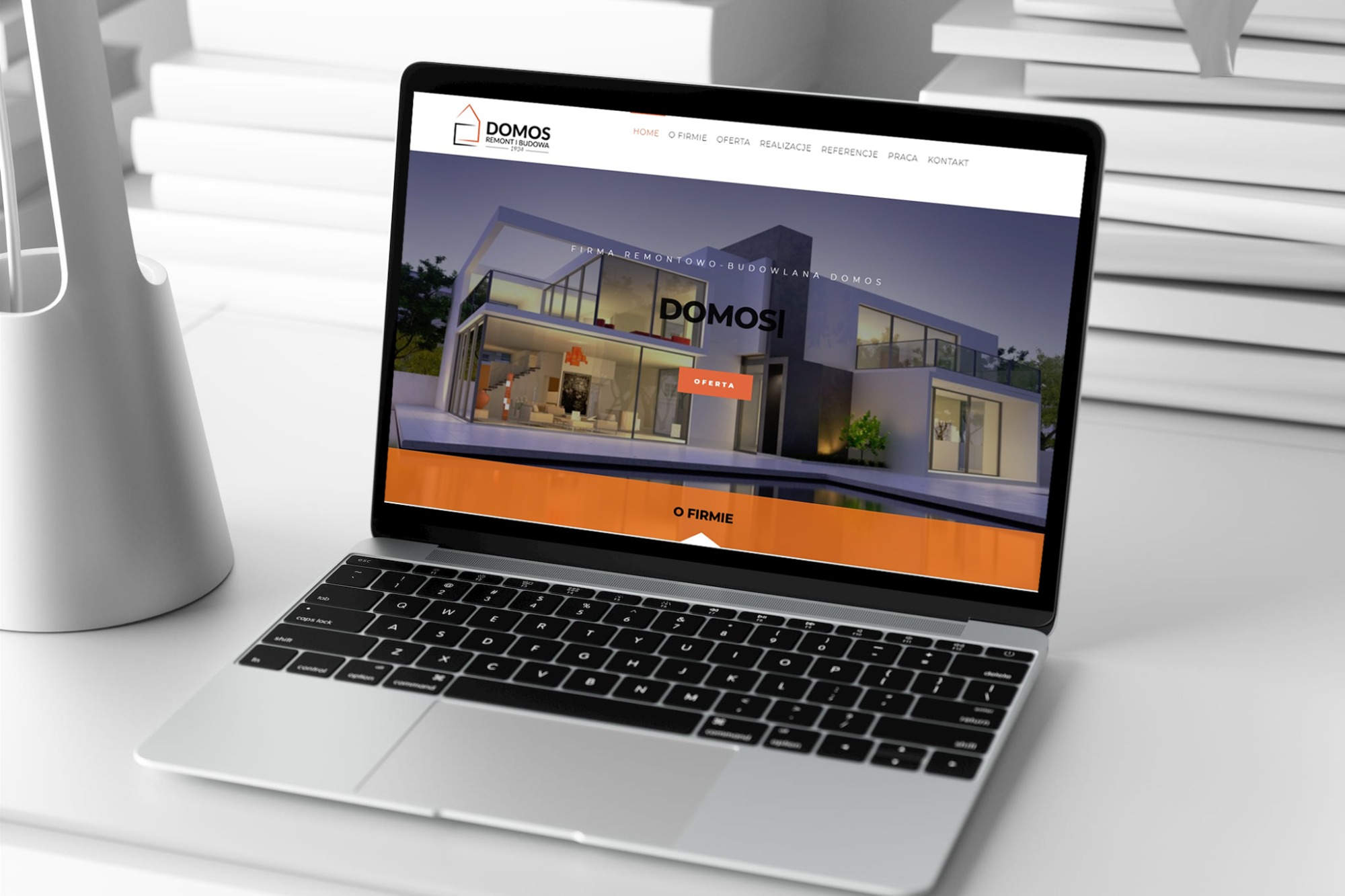 Laptop wyświetla stronę internetową firmy remontowo-budowlanej Domos, z wizualizacją nowoczesnego domu jednorodzinnego, na białym biurku obok lampy.