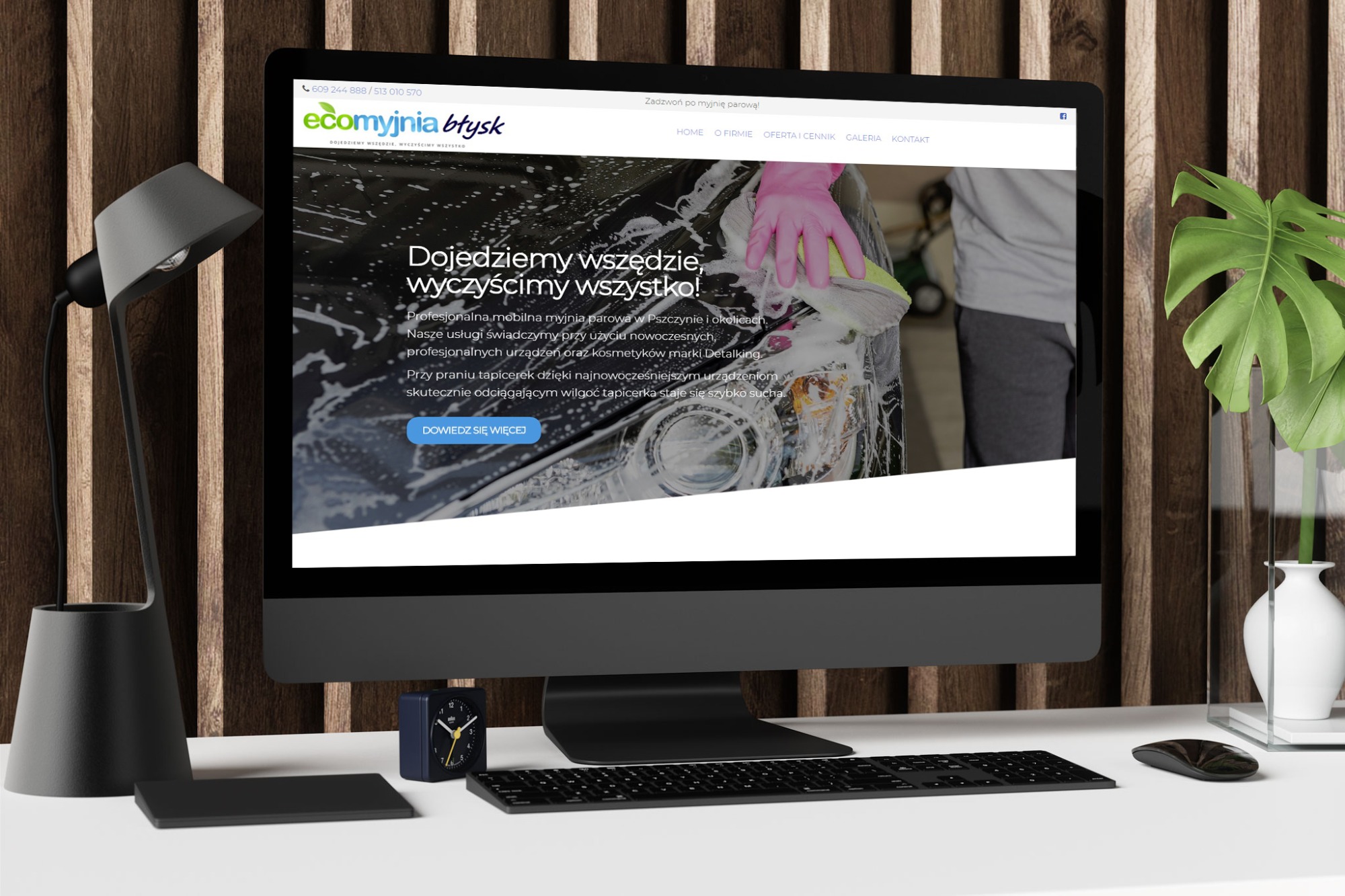 Monitor wyświetla stronę internetową Ecomyjnia Błysk, prezentującą mycie samochodu z użyciem piany i rękawicy, na biurku z lampką, zegarem i klawiaturą.