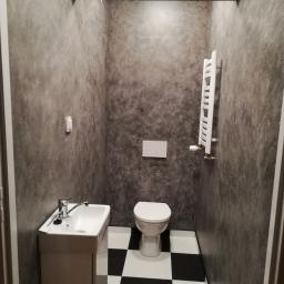 Usługi remontowo-budowlane Łukasz ziemian - Wnętrze małej łazienki z umywalką na szafce, toaletą i grzejnikiem drabinkowym, ściany wykończone w szarym, nieregularnym wzorze, podłoga w czarno-białą szachownicę.