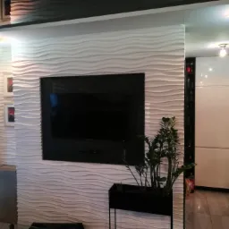 Telewizor zawieszony na ścianie z dekoracyjnymi panelami 3D w kształcie fal, obok okno z żaluzjami i minimalistyczna kuchnia w tle.