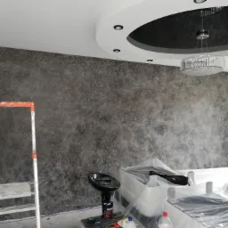 Pokój po wykonaniu dekoracyjnej ściany w odcieniach szarości, zabezpieczone meble folią, widoczna drabina malarska i lampa wisząca z kryształkami.