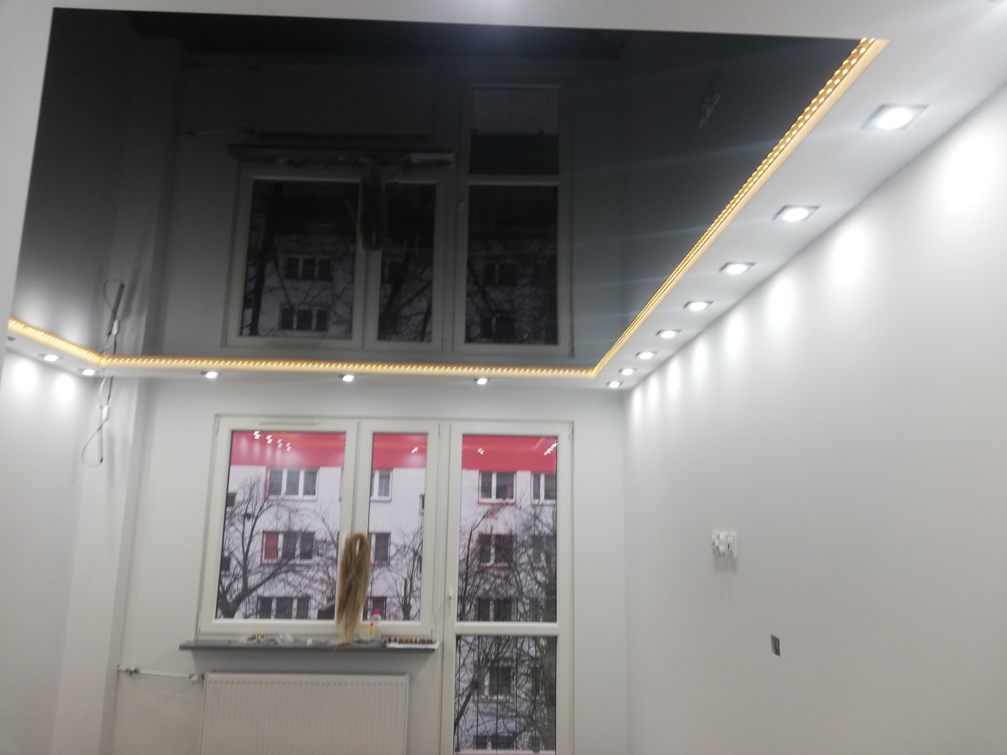 Czarny, błyszczący sufit napinany z oświetleniem LED wzdłuż krawędzi w pokoju z białymi ścianami i oknami.