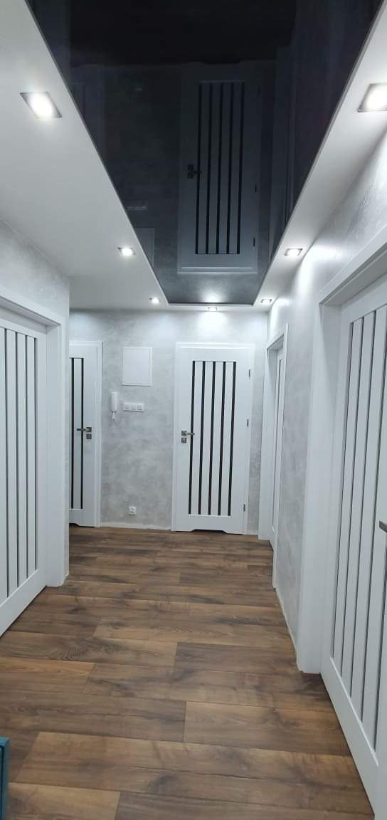 Korytarz z ciemną, lustrzaną powierzchnią sufitu, wpuszczanymi lampami, szarą ścianą z efektem dekoracyjnym i brązowymi panelami podłogowymi oraz białymi drzwiami z czarnymi wstawkami.