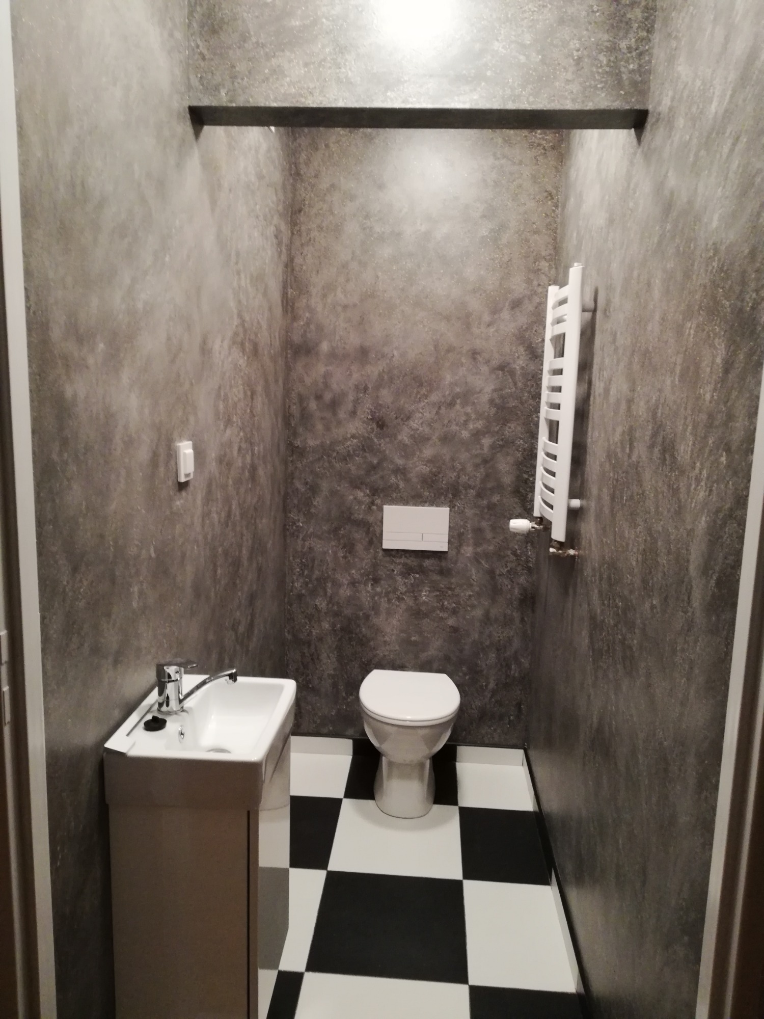 Wnętrze małej łazienki z umywalką na szafce, toaletą i grzejnikiem drabinkowym, ściany wykończone w szarym, nieregularnym wzorze, podłoga w czarno-białą szachownicę.