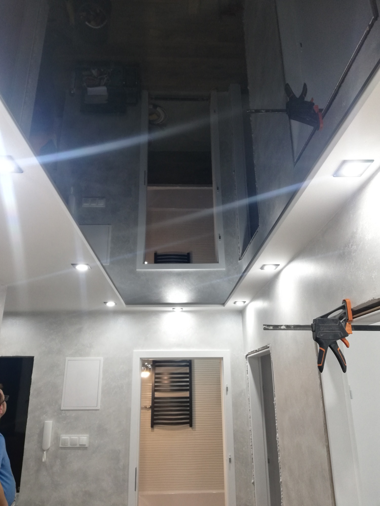 Błyszczący sufit z zamontowanymi lampami i otworem rewizyjnym, widoczne ślady szpachlowania na ścianach, w tle widoczne drzwi do łazienki z grzejnikiem.