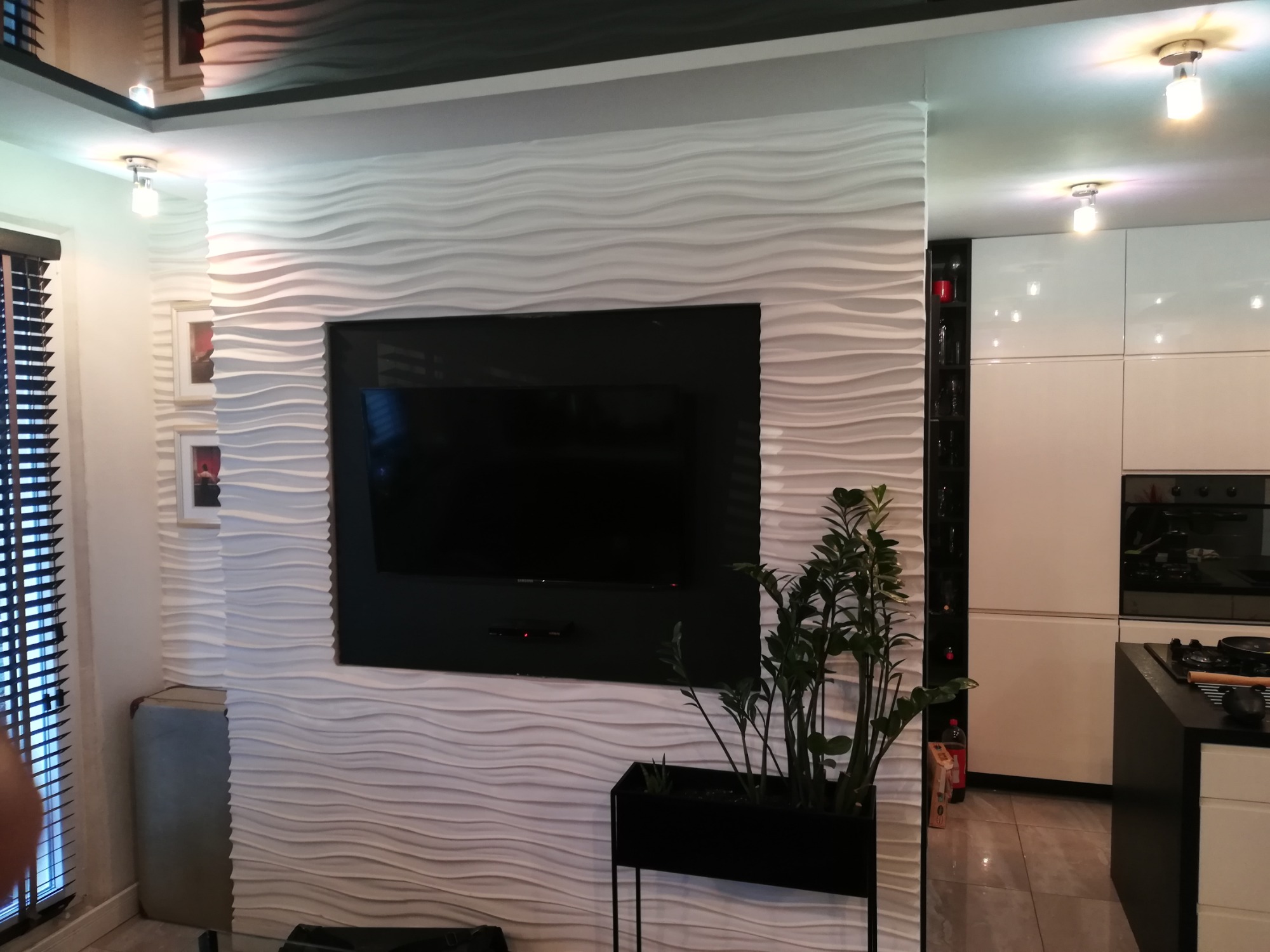 Telewizor zawieszony na ścianie z dekoracyjnymi panelami 3D w kształcie fal, obok okno z żaluzjami i minimalistyczna kuchnia w tle.