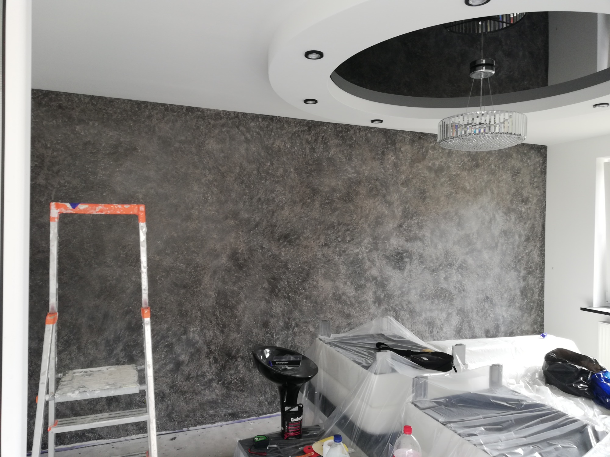 Pokój po wykonaniu dekoracyjnej ściany w odcieniach szarości, zabezpieczone meble folią, widoczna drabina malarska i lampa wisząca z kryształkami.