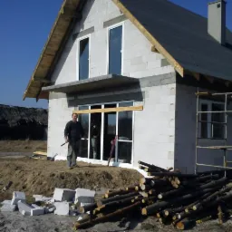 Budowa domu z białych bloczków: widoczne ściany, okna, dach w trakcie prac. Na pierwszym planie stos drewna i pracownik z narzędziem. W tle budynek gospodarczy.