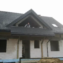 Dom w stanie surowym otwartym z ciemną dachówką, widoczny pracownik na dachu. Budynek z białego bloczka, okna bez szyb, teren budowy.