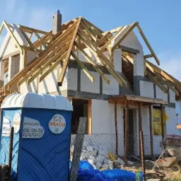 Budowa domu jednorodzinnego w trakcie realizacji, widoczna konstrukcja dachu, ściany z pustaków, przenośna toaleta budowlana na pierwszym planie, bałagan na placu budowy.