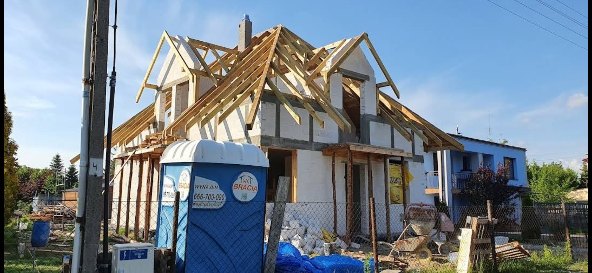 Budowa domu jednorodzinnego w trakcie realizacji, widoczna konstrukcja dachu, ściany z pustaków, przenośna toaleta budowlana na pierwszym planie, bałagan na placu budowy.