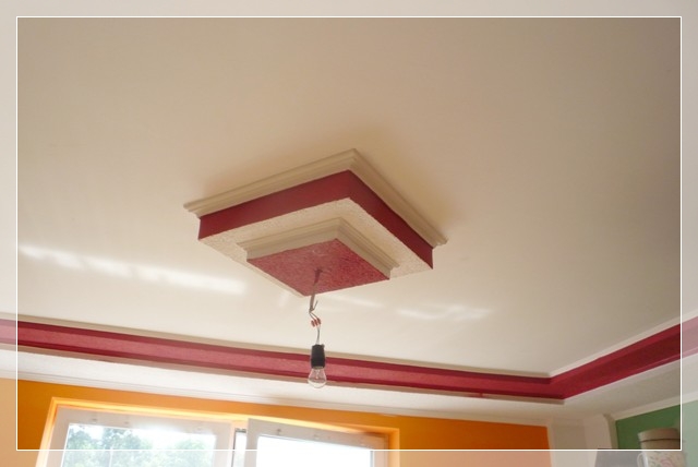 Wykończony sufit z dekoracyjnym, kaskadowym elementem centralnym w kolorze czerwonym i beżowym, z wiszącą żarówką na kablu, oraz czerwonym paskiem dekoracyjnym wzdłuż ścian.