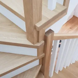 Jasne, dębowe stopnie schodów spiralnych z białymi podstopnicami i balustradą, widok z góry na fragment konstrukcji.