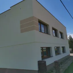 Nowoczesny budynek jednorodzinny z jasną elewacją, akcentami w postaci poziomych pasów w odcieniach beżu i brązowymi ramami okiennymi, sfotografowany w słoneczny dzień z widocznym błękitnym niebem.