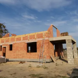Surowy stan budynku jednorodzinnego z czerwonej cegły, widoczne otwory okienne i drzwiowe, zadaszenie tarasu wsparte na betonowych słupach, drabina oparta o ścianę, teren budowy.
