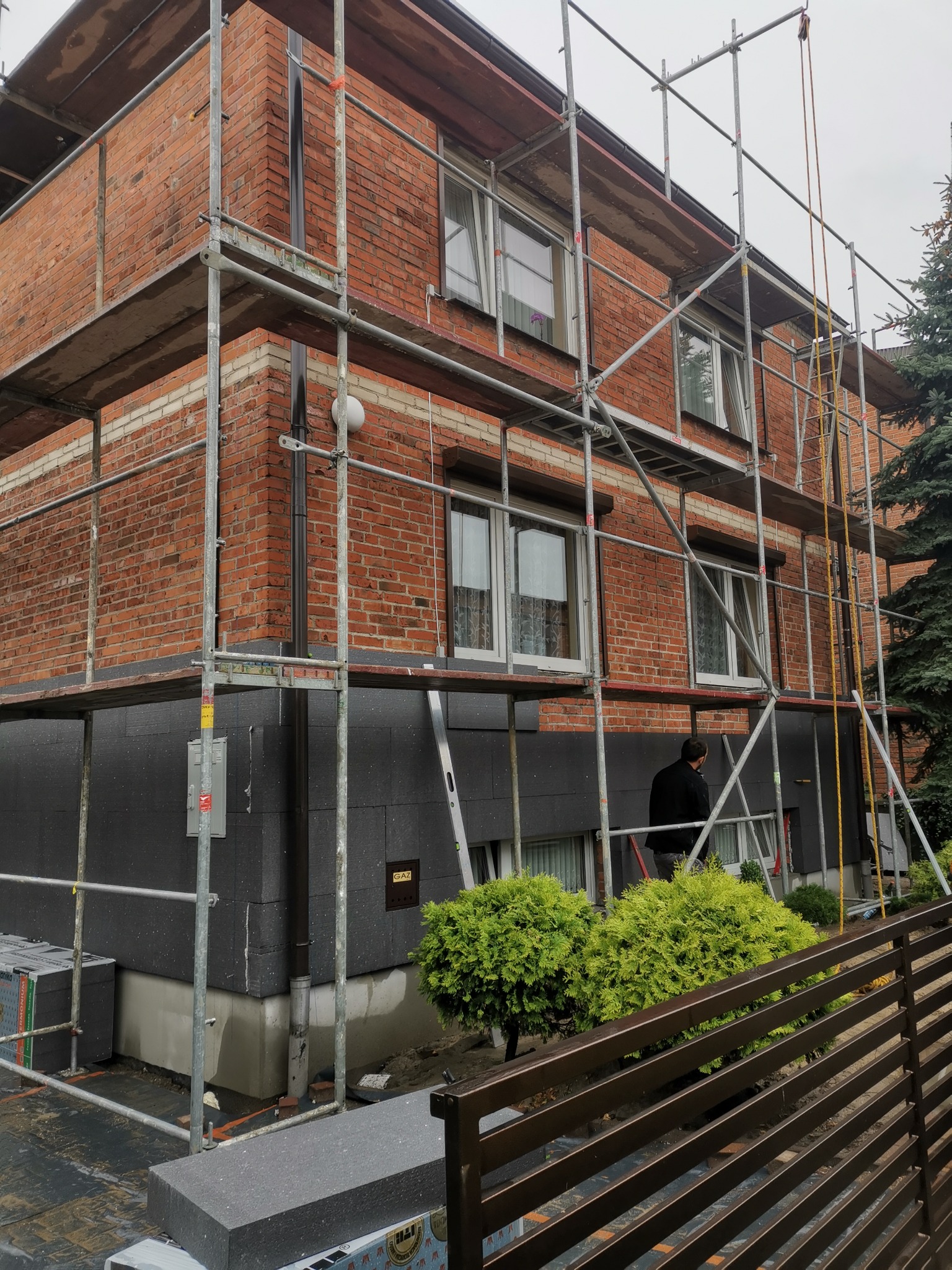 Dwupiętrowy dom z czerwonej cegły w trakcie ocieplania, widoczne szare płyty izolacyjne na dolnej części elewacji, rusztowania wokół budynku, osoba w ciemnej kurtce w tle.