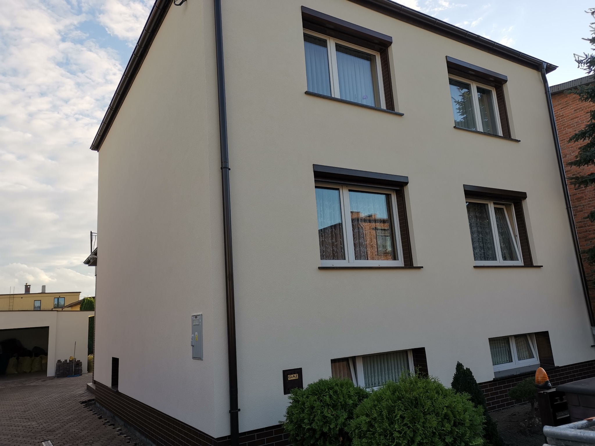 Dwupiętrowy dom po renowacji elewacji, z jasną, gładką powierzchnią, ciemnymi ramami okiennymi i rynnami, widoczne krzewy i kostka brukowa.