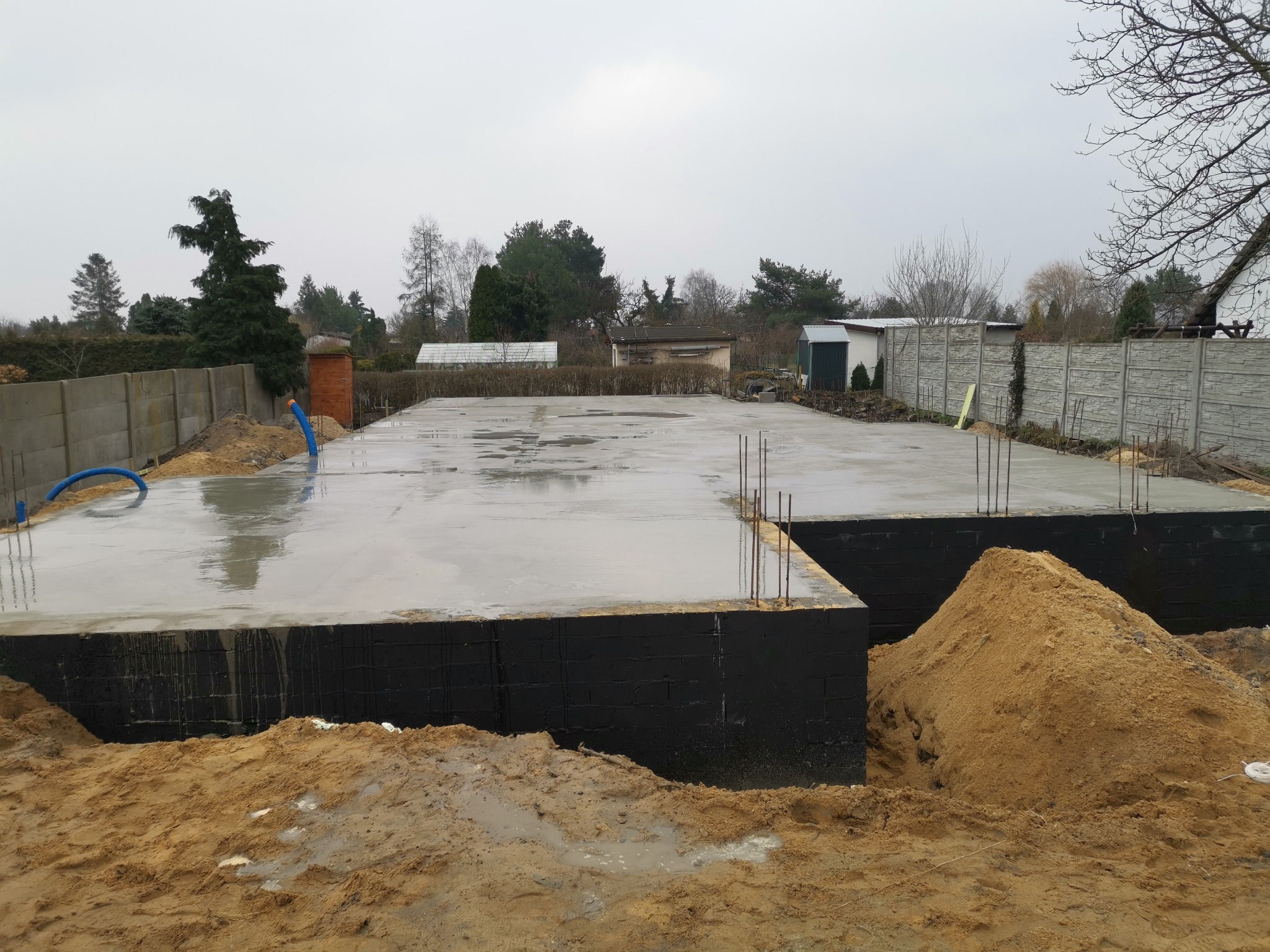 Wylana, mokra płyta fundamentowa na gruncie z wystającymi prętami zbrojeniowymi, zabezpieczona czarną izolacją, z widocznym nasypem piasku.