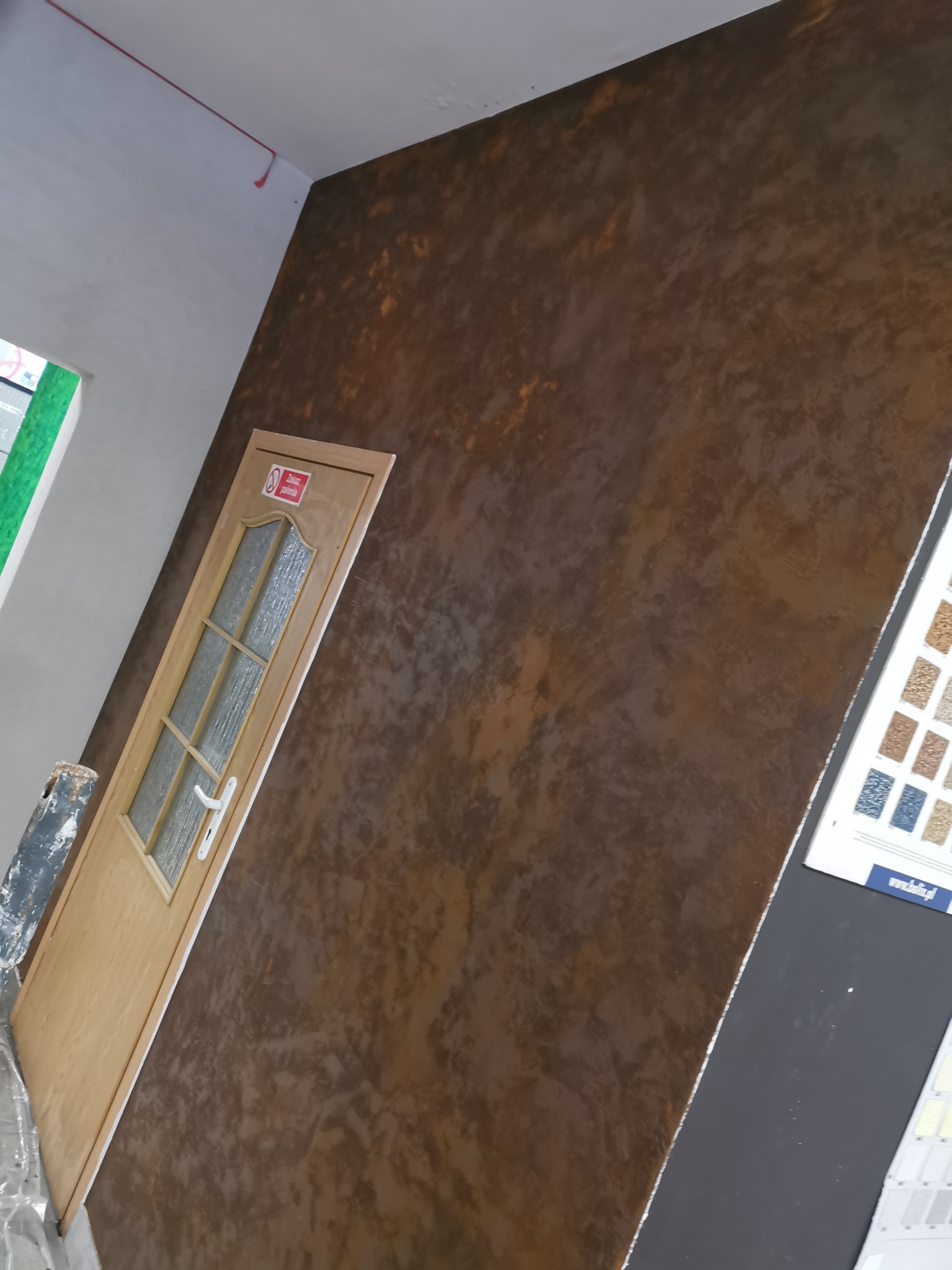 Ściana wewnętrzna pokryta dekoracyjnym tynkiem w odcieniach brązu, obok drewniane drzwi z przeszkleniami i próbnik kolorów na czarnym tle.