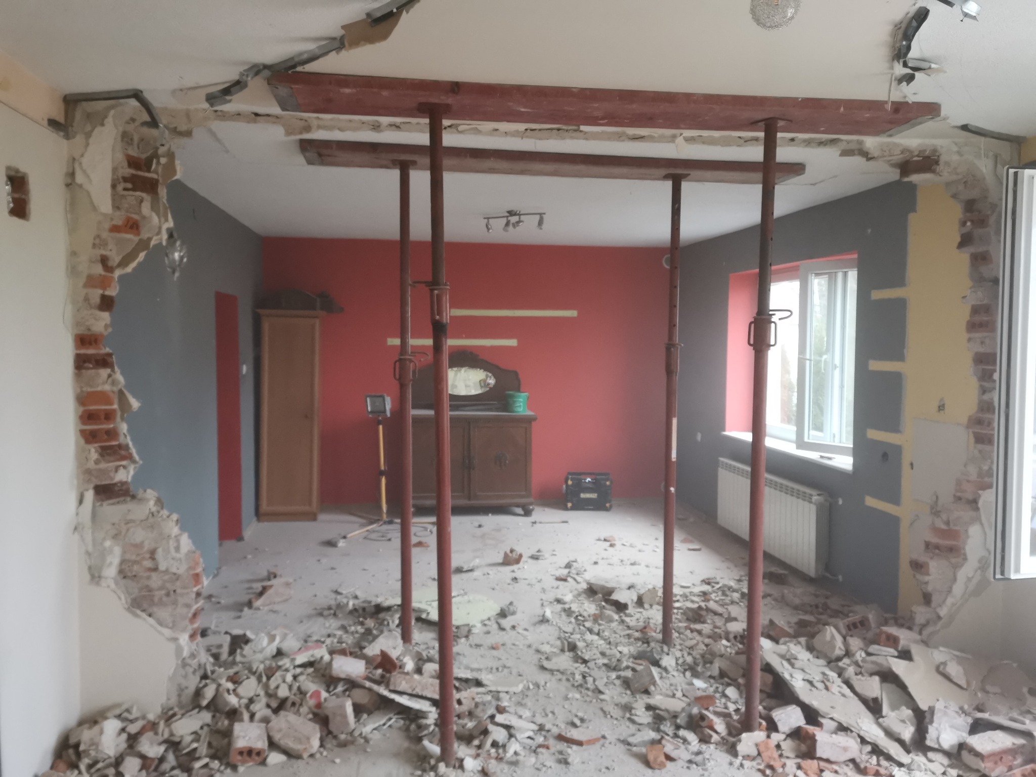 Wnętrze w trakcie remontu: usunięte fragmenty ścian odsłaniające cegły, metalowe podpory podtrzymujące sufit, gruz na podłodze, widoczne meble w tle.