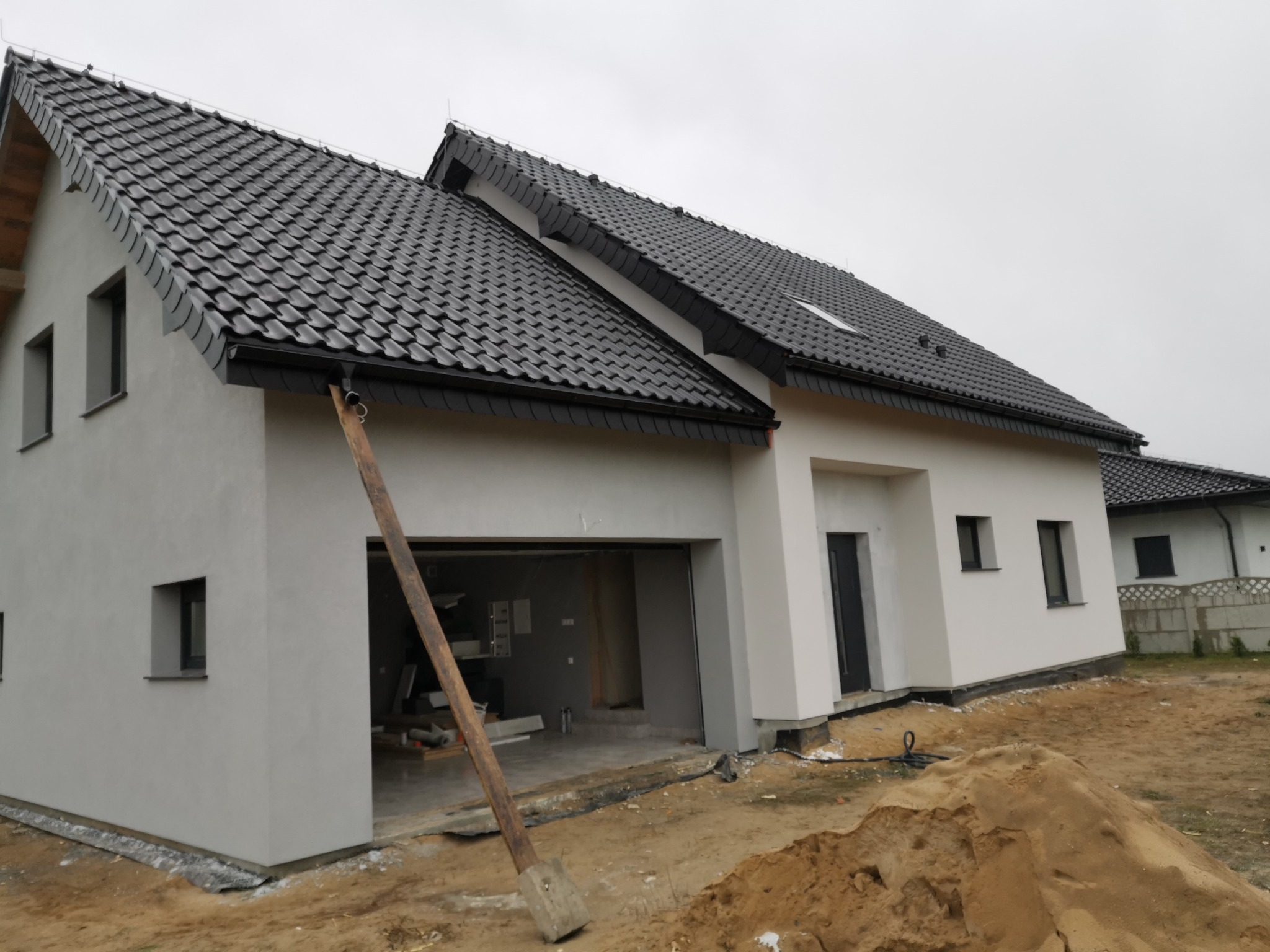 Nowoczesny dom jednorodzinny z szarą elewacją i czarną dachówką, widoczna sterta piasku i drewniana podpora, sugerująca prace wykończeniowe.