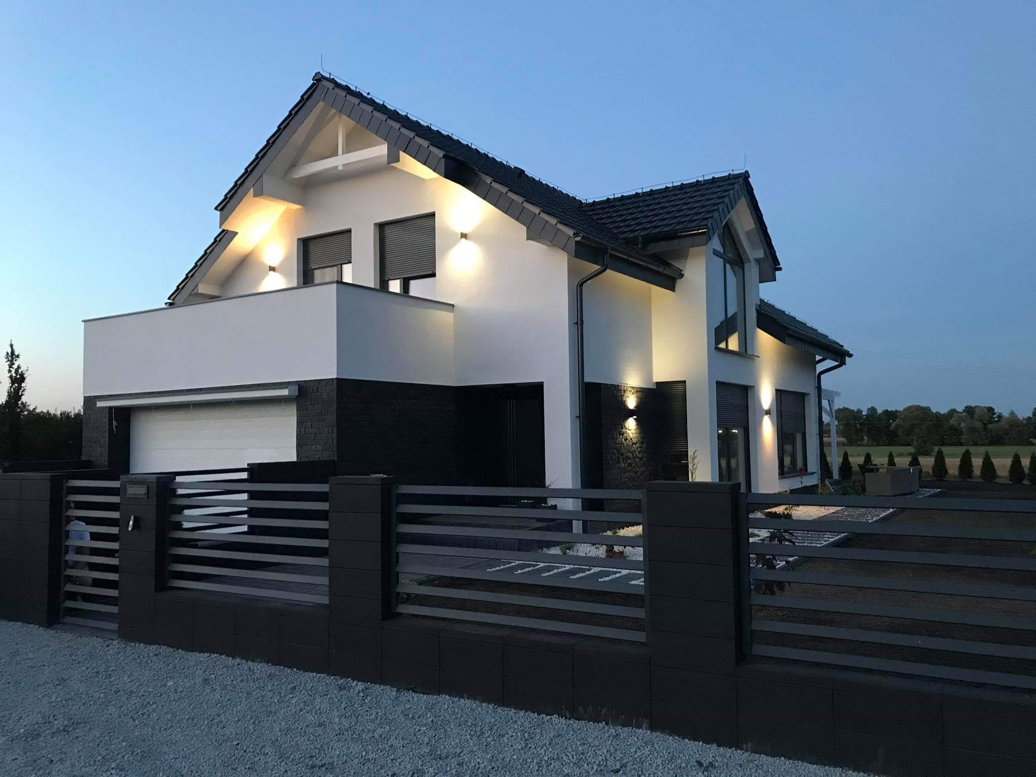 Nowoczesny dom jednorodzinny z białymi ścianami i czarną elewacją, oświetlony zewnętrznymi lampami, z czarnym ogrodzeniem panelowym na tle wieczornego nieba.