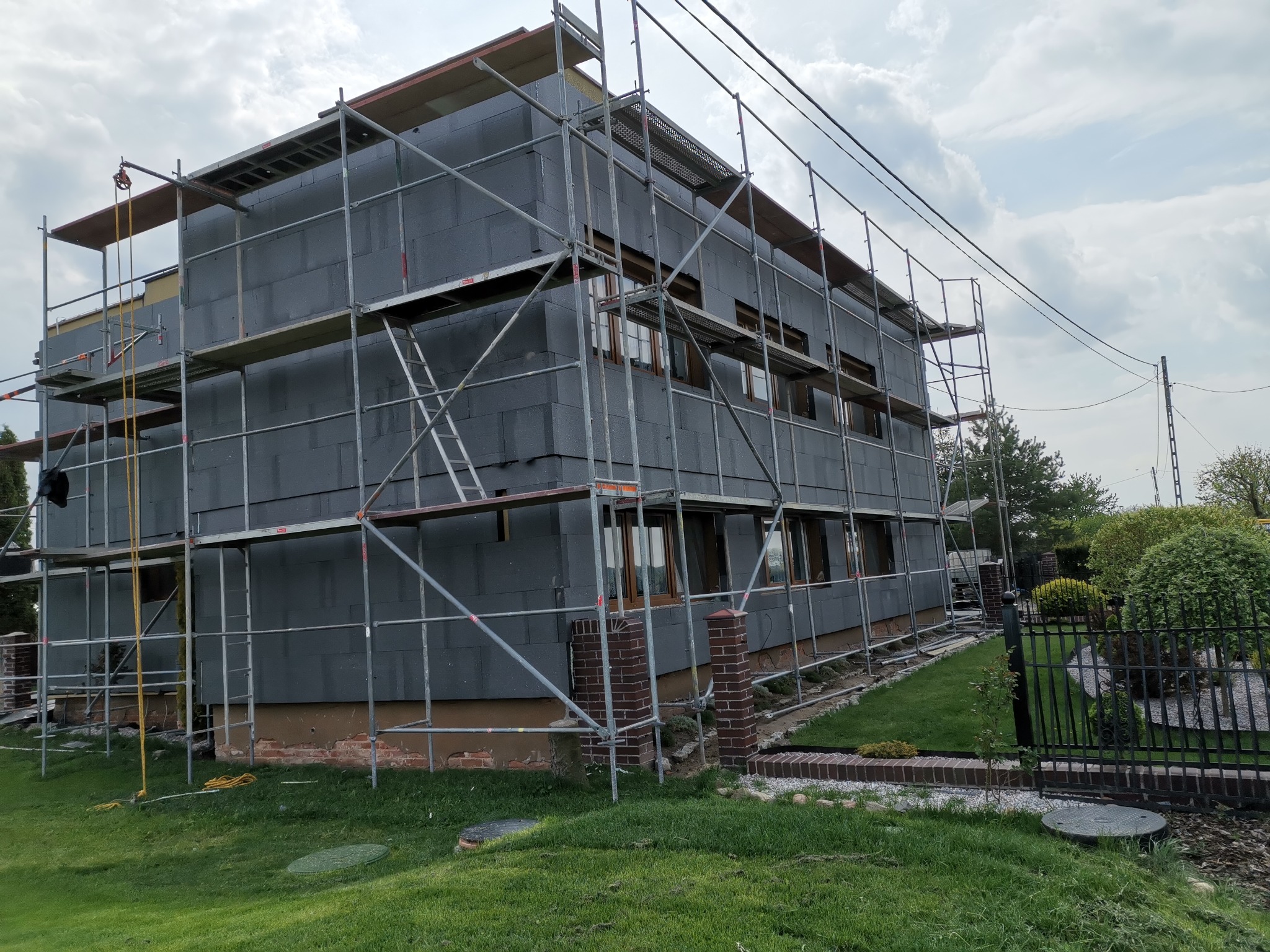 Dwukondygnacyjny dom w trakcie prac izolacyjnych, pokryty szarymi płytami styropianowymi, otoczony rusztowaniem; widoczne fragmenty elewacji z cegły oraz zadbany trawnik.