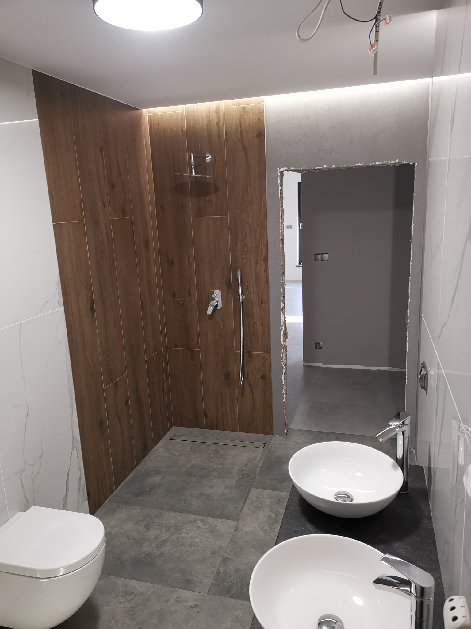 Nowoczesna łazienka z dwoma umywalkami nablatowymi, prysznicem typu walk-in z drewnopodobnymi płytkami i szarymi płytkami podłogowymi, widok na sąsiednie pomieszczenie przez otwarte drzwi.