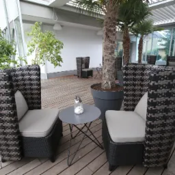 Zadaszony taras z drewnianą podłogą, wyposażony w dwa wysokie, plecione fotele z szarymi poduszkami, mały okrągły stolik z dekoracją w szklanym lampionie oraz donice z palmami.