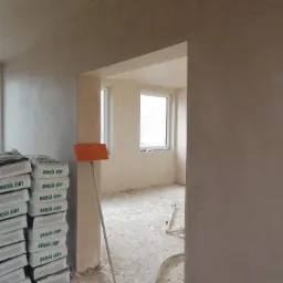 Świeżo położone, jasne tynki na ścianach wewnątrz budynku, widoczne worki z zaprawą Dolina Nidy i pomarańczowy pędzel do rozprowadzania tynku.