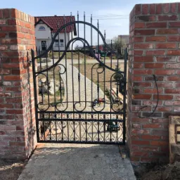 Ozdobna, czarna brama z kutego żelaza, osadzona między dwoma murowanymi filarami z czerwonej cegły, widok na kamienną ścieżkę i dom w tle, narzędzia budowlane leżące na ziemi.