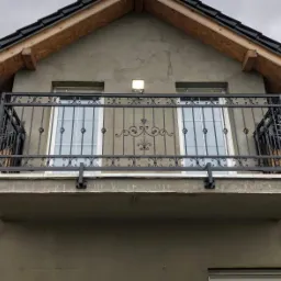 Czarna, dekoracyjna balustrada balkonowa z kutymi elementami, zamontowana na szarym budynku z widocznymi oknami i czarnym dachem.