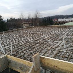 Odbierajznami - Zbrojenie stropu z widocznymi elementami szalunku drewnianego i wystającymi rurami instalacyjnymi, w tle zabudowa jednorodzinna i drzewa.