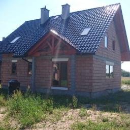 Odbierajznami - Budynek jednorodzinny w stanie surowym otwartym z ciemną dachówką, widoczne okna z drewnianą stolarką i elementy konstrukcji dachu z czerwonego drewna, otoczony wysoką trawą i polnymi kwiatami.