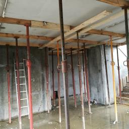 Odbierajznami - Wnętrze surowego budynku w trakcie budowy ze stalowymi podporami stropowymi i drewnianymi belkami podtrzymującymi szalunek, częściowo zalane wodą, z drabiną opartą o ścianę.