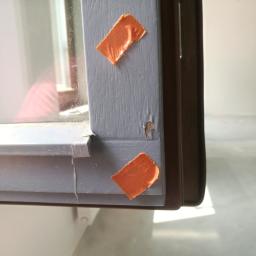 Odbierajznami - Szczegółowe ujęcie uszkodzonego, szarego ramy okiennej z widocznym ubytkiem w drewnie i dwoma kawałkami pomarańczowej taśmy maskującej, prawdopodobnie przed malowaniem lub naprawą.