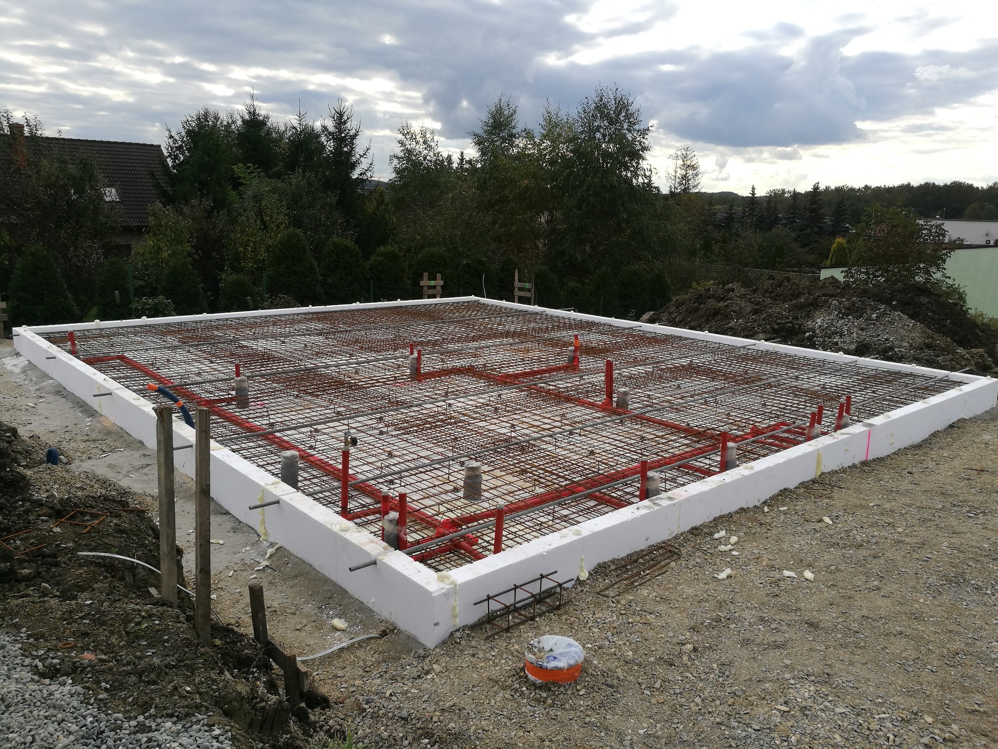 Przygotowanie płyty fundamentowej z izolacją termiczną, zbrojenie stalowe siatką i rurami instalacyjnymi w kolorze czerwonym, widok z góry na placu budowy.