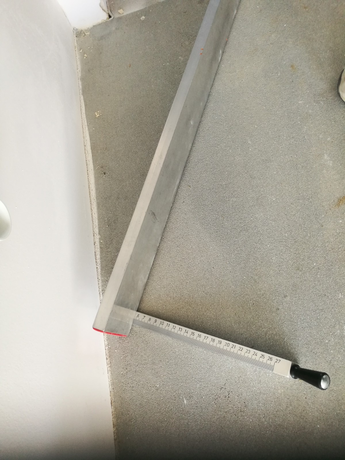 Pomiar kąta prostego przy ścianie za pomocą kątownika z podziałką centymetrową na szarej, chropowatej powierzchni.