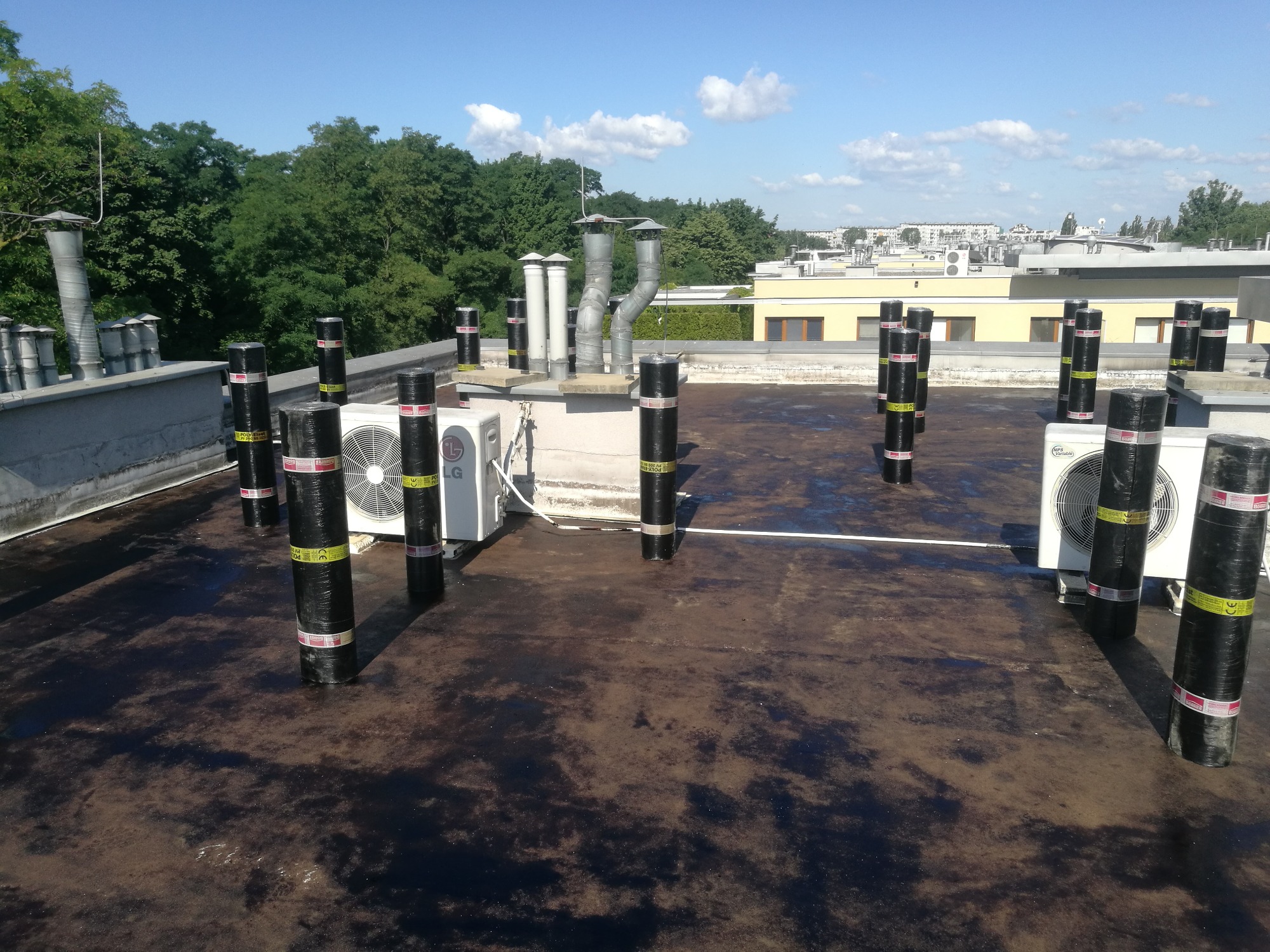 Wykonanie hydroizolacji dachu płaskiego z papy termozgrzewalnej, widoczne kominy wentylacyjne, klimatyzatory i zabezpieczone rury na dachu budynku.