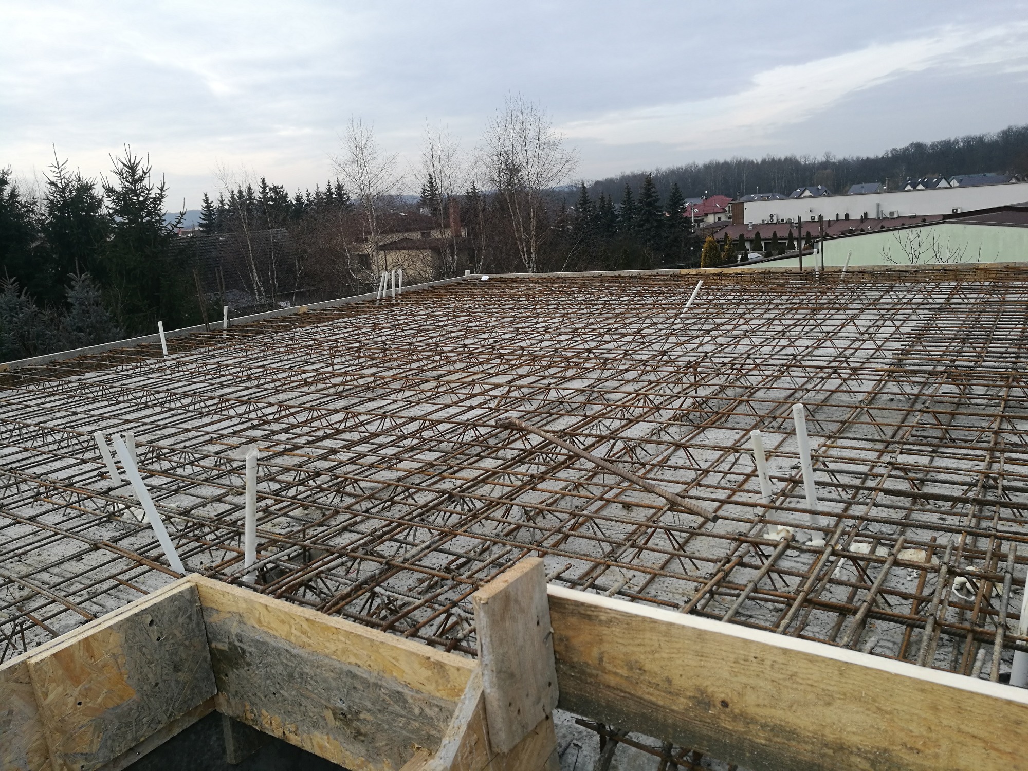 Zbrojenie stropu z widocznymi elementami szalunku drewnianego i wystającymi rurami instalacyjnymi, w tle zabudowa jednorodzinna i drzewa.