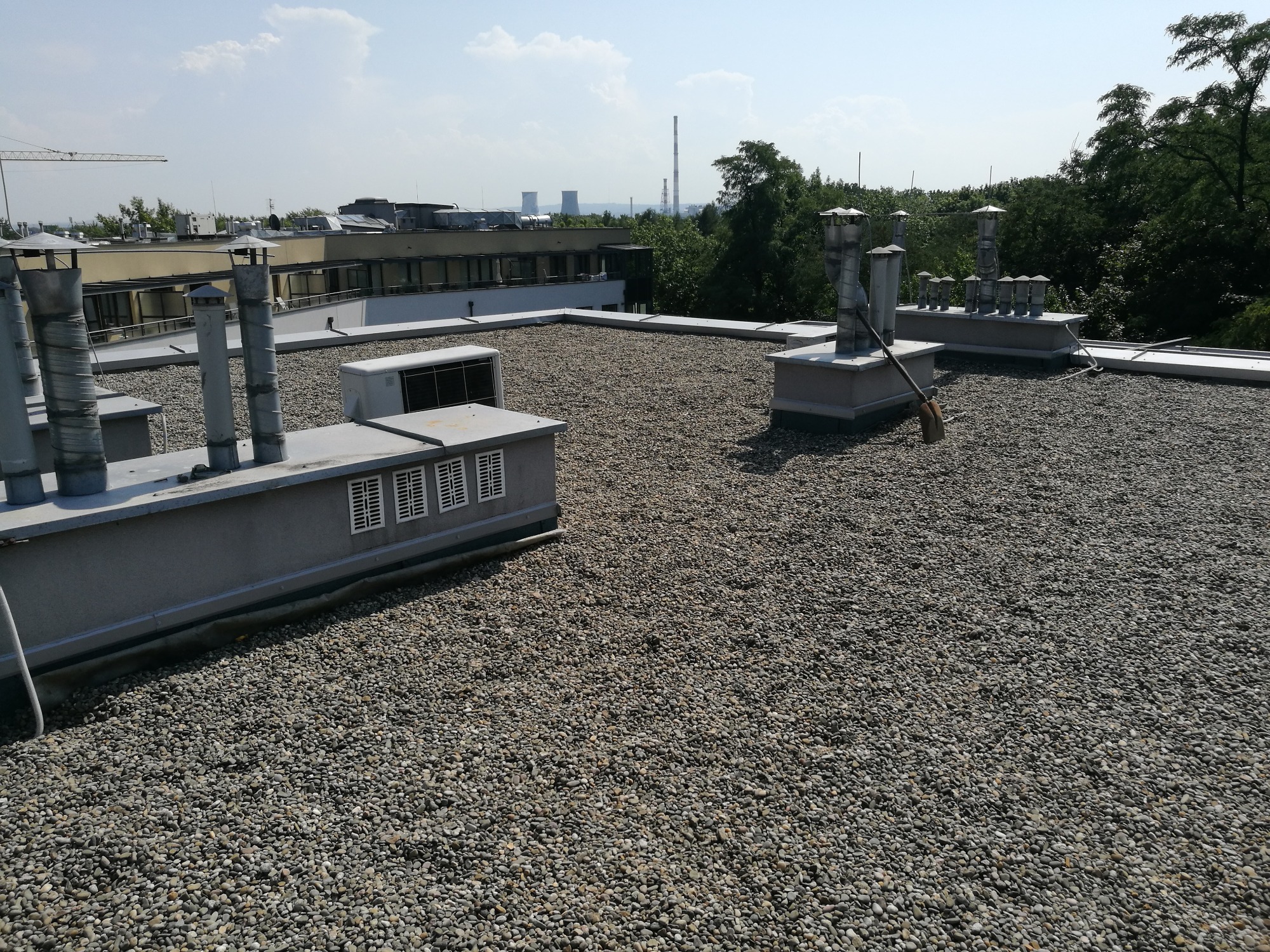 Płaski dach budynku pokryty drobnym żwirem, widoczne kominy wentylacyjne, klimatyzatory i budynki w tle z kominami przemysłowymi.