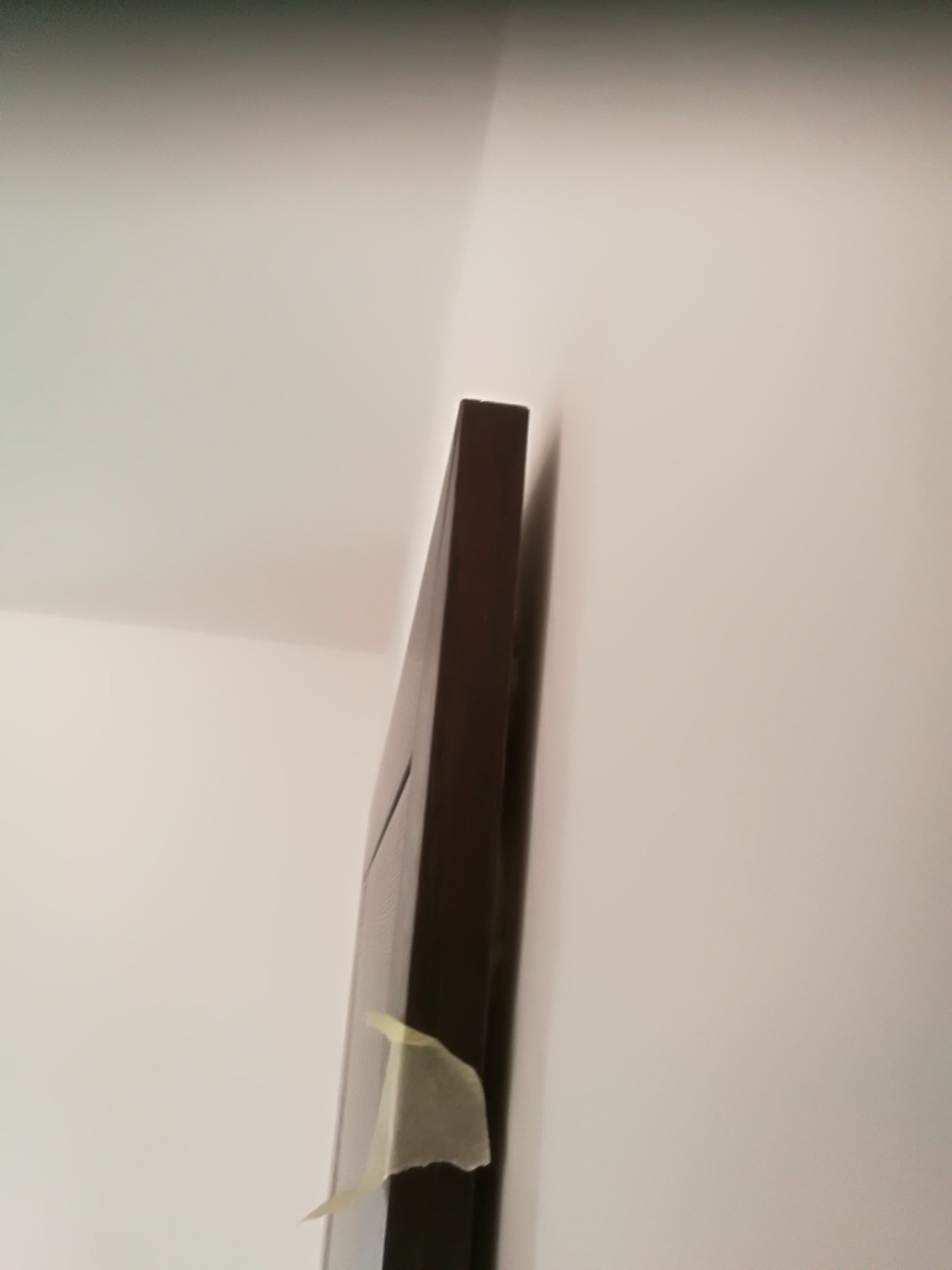 Boczny widok ciemnej, drewnianej ramy drzwiowej przyklejonej do białej ściany za pomocą żółtej taśmy malarskiej, widoczne niedoskonałości i nierówności powierzchni.