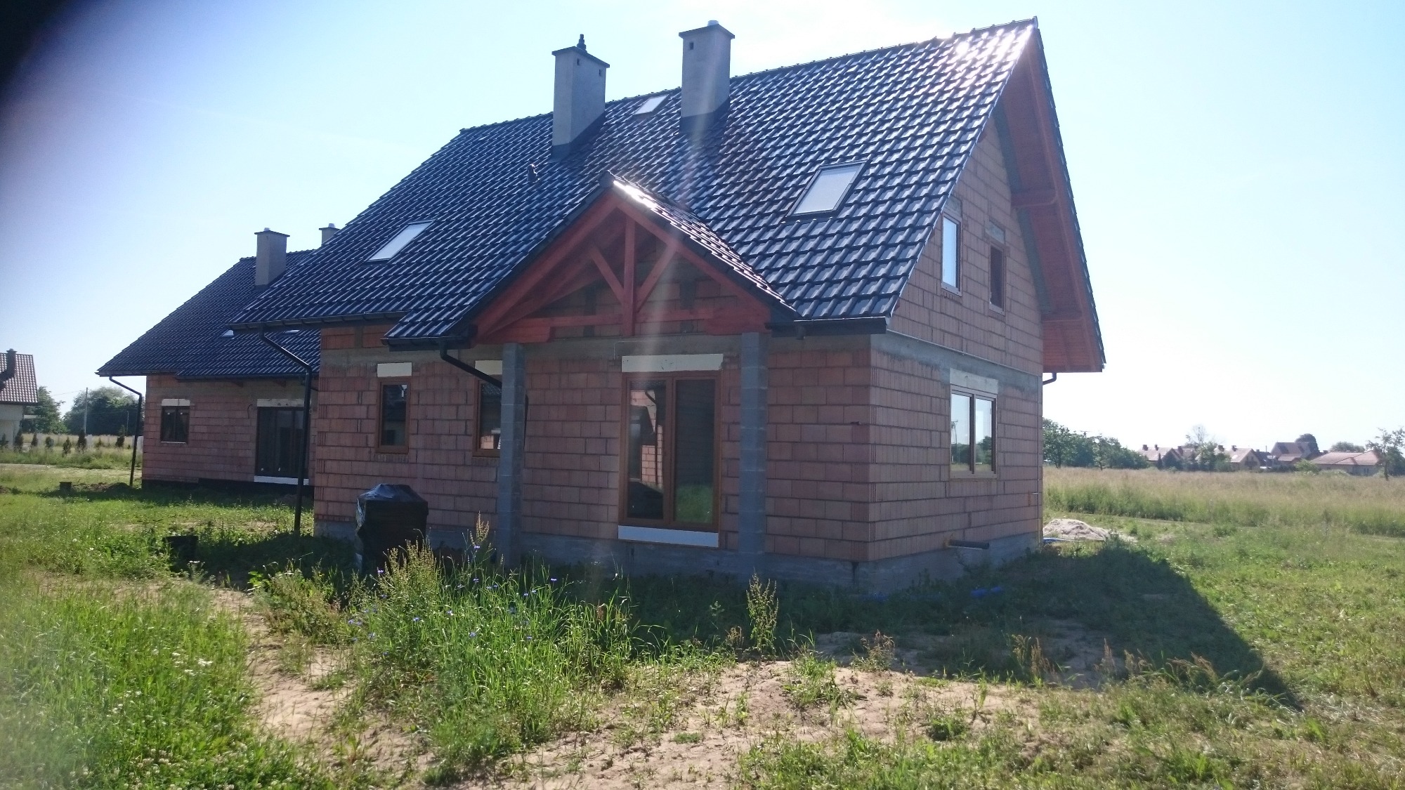 Budynek jednorodzinny w stanie surowym otwartym z ciemną dachówką, widoczne okna z drewnianą stolarką i elementy konstrukcji dachu z czerwonego drewna, otoczony wysoką trawą i polnymi kwiatami.