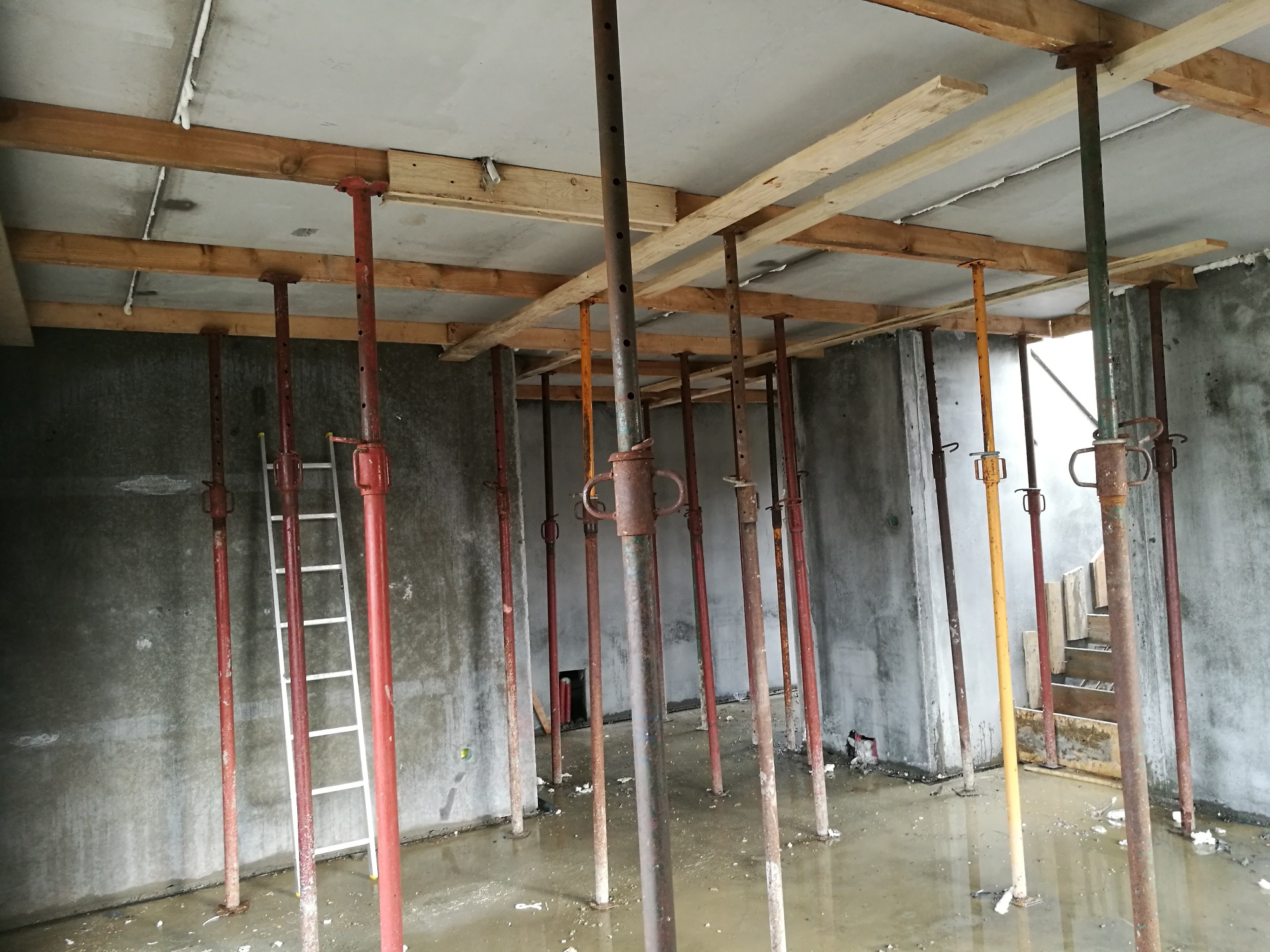 Wnętrze surowego budynku w trakcie budowy ze stalowymi podporami stropowymi i drewnianymi belkami podtrzymującymi szalunek, częściowo zalane wodą, z drabiną opartą o ścianę.