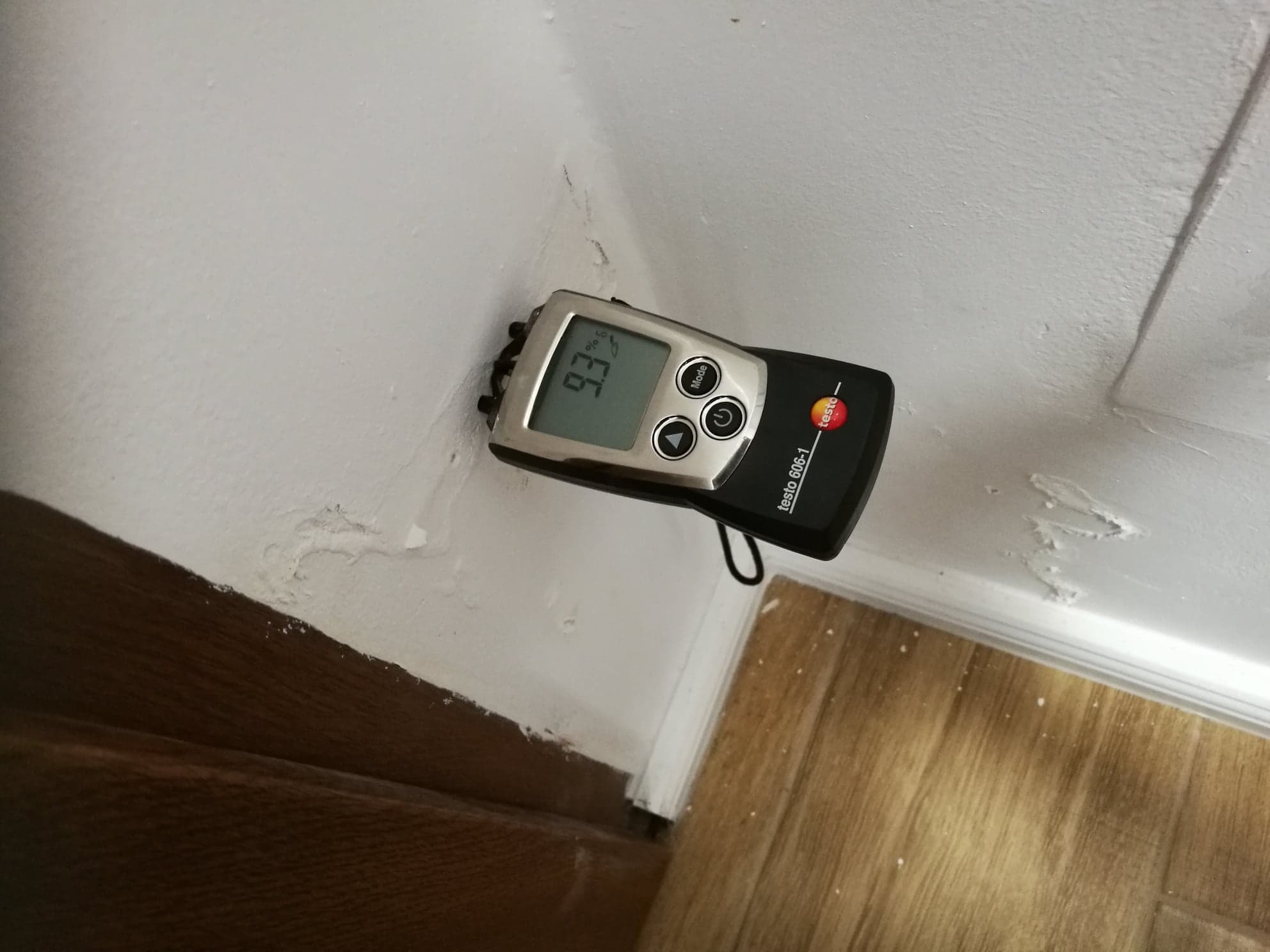 Pomiar wilgotności ściany przy podłodze za pomocą wilgotnościomierza Testo 606-1, wskazującego 9.3%, z widocznymi śladami zawilgocenia i odchodzącej farby.