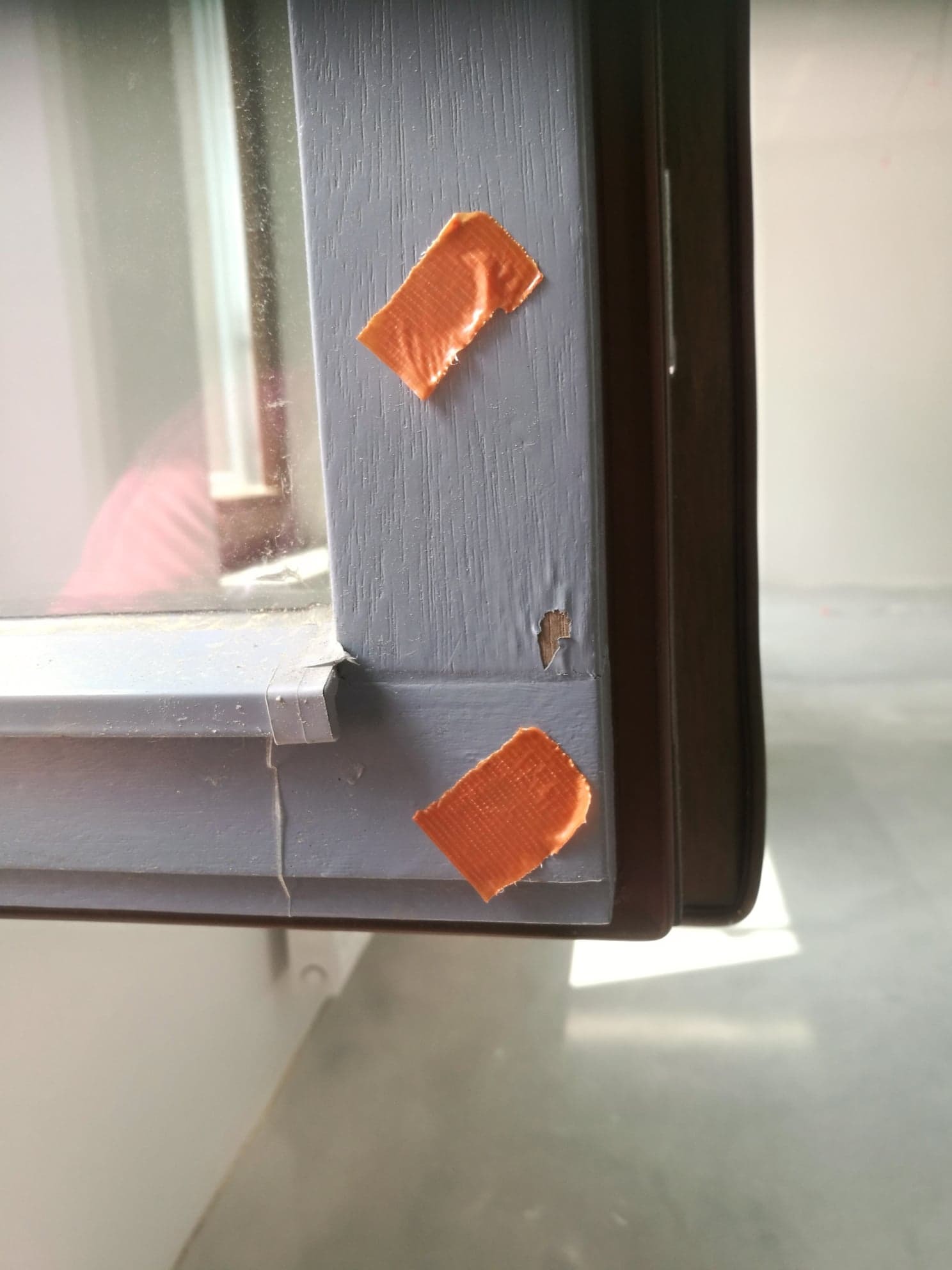 Szczegółowe ujęcie uszkodzonego, szarego ramy okiennej z widocznym ubytkiem w drewnie i dwoma kawałkami pomarańczowej taśmy maskującej, prawdopodobnie przed malowaniem lub naprawą.